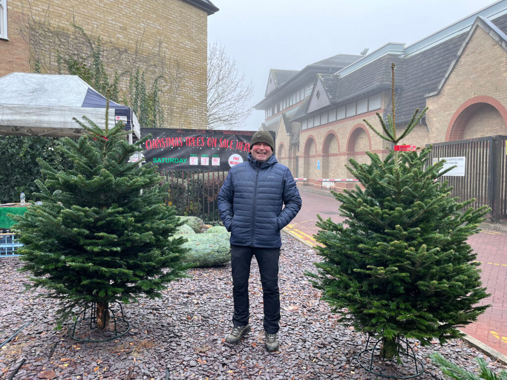 A man standing outside in between two Christmas Trees.