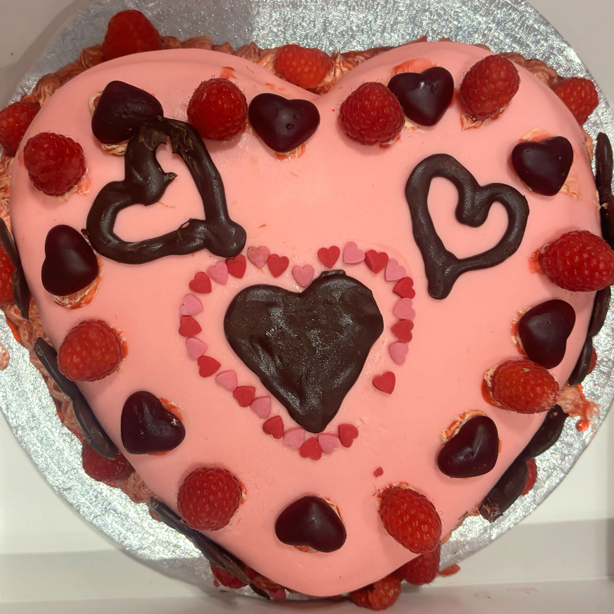 a pink and red cake in the shape of a heart with chocolate hearts and strawberries around the edge for the ebs cake competition